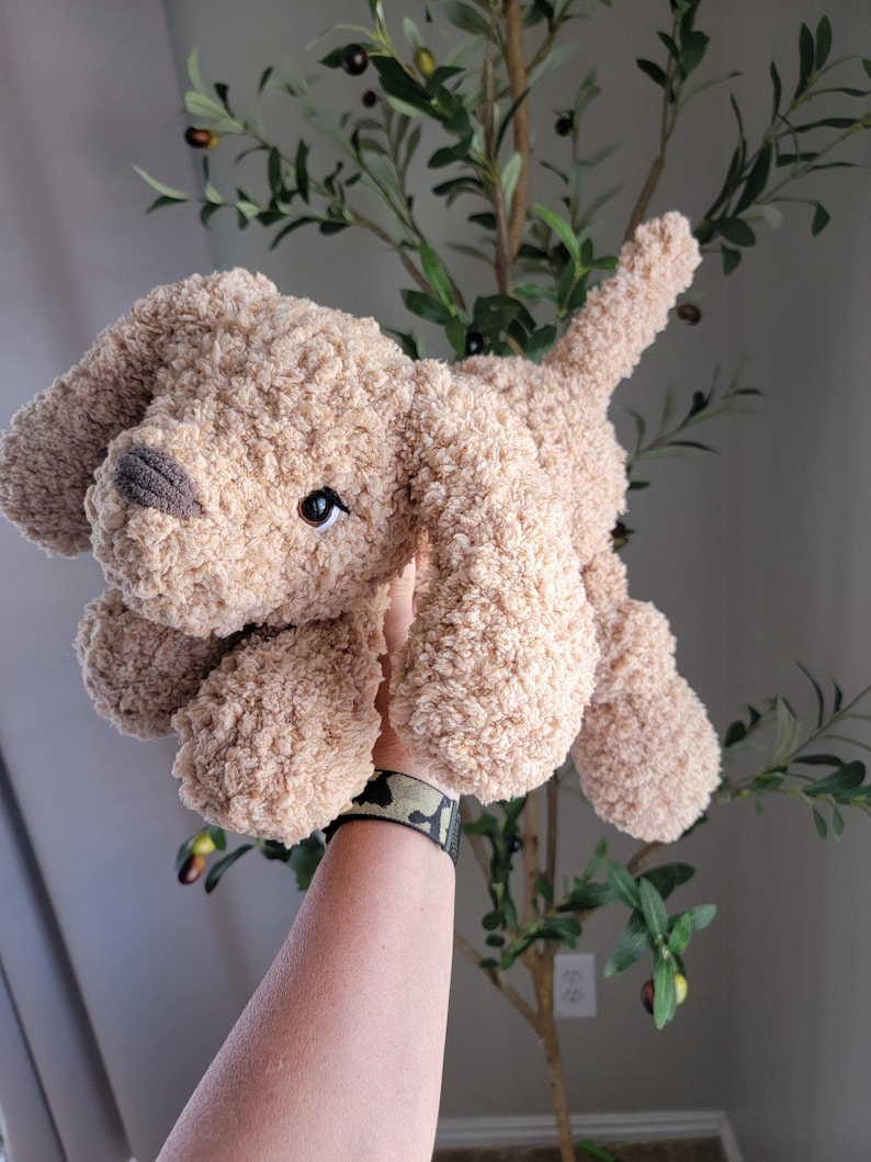 Puede incluir: Un juguete de peluche de perro de color marr&oacute;n claro con textura esponjosa. El perro est&aacute; sentado con las patas extendidas y la cabeza inclinada hacia un lado.