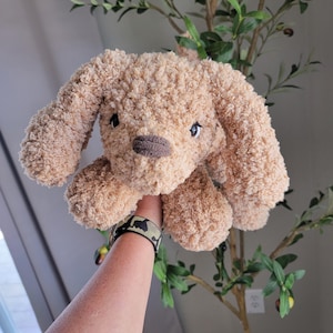 Puede incluir: Un juguete de peluche de color marr&oacute;n claro en forma de perro con ojos negros y nariz marr&oacute;n. El perro est&aacute; siendo sostenido por una mano que lleva una pulsera de camuflaje.
