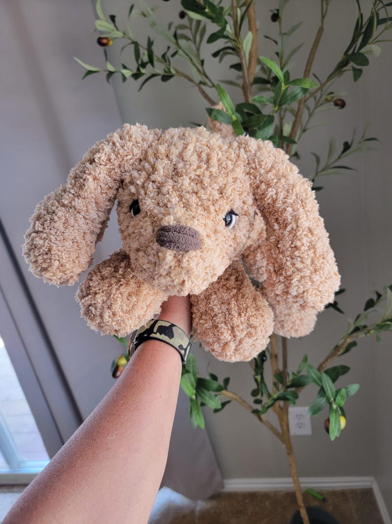 Puede incluir: Un juguete de peluche de color marr&oacute;n claro en forma de perro con ojos negros y nariz marr&oacute;n. El perro est&aacute; siendo sostenido por una mano que lleva una pulsera de camuflaje.