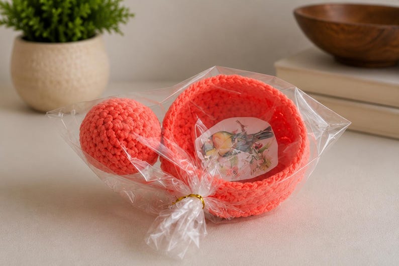 May include: A bright neon orange crocheted bowl and a matching crocheted sphere packaged together in a clear plastic bag. Inside the bowl is a small, rectangular paper insert featuring a detailed illustration of a bird perched on a branch.