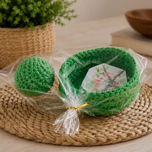 May include: A bright green crocheted bowl and a matching green crocheted sphere packaged together in a clear plastic bag with a gold twist tie. Inside the bowl is a small hexagonal card featuring a printed illustration of pink cherry blossoms.