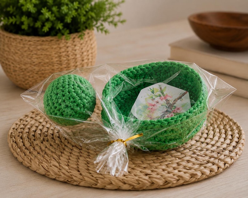May include: A bright green crocheted bowl and a matching green crocheted sphere packaged together in a clear plastic bag with a gold twist tie. Inside the bowl is a small hexagonal card featuring a printed illustration of pink cherry blossoms.