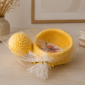 May include: A bright yellow crocheted bowl and a matching yellow crocheted sphere packaged together in clear plastic wrap with a gold twist tie. A small rectangular tag with a floral print is visible inside the yellow crocheted bowl.