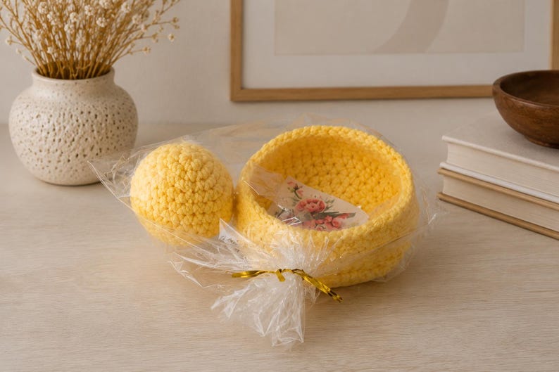 May include: A bright yellow crocheted bowl and a matching yellow crocheted sphere packaged together in clear plastic wrap with a gold twist tie. A small rectangular tag with a floral print is visible inside the yellow crocheted bowl.