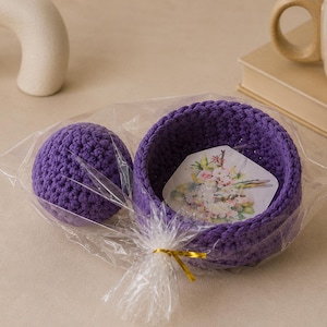 May include: A purple crocheted bowl and a matching purple crocheted sphere packaged together in a clear plastic bag with a gold twist tie. Inside the bowl is a hexagonal card featuring a colorful floral illustration with pink and white flowers.