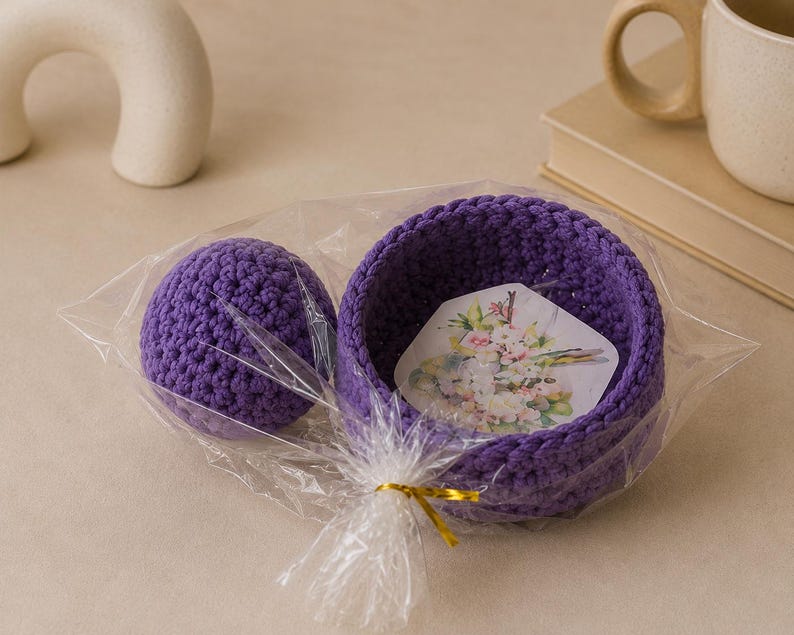 May include: A purple crocheted bowl and a matching purple crocheted sphere packaged together in a clear plastic bag with a gold twist tie. Inside the bowl is a hexagonal card featuring a colorful floral illustration with pink and white flowers.