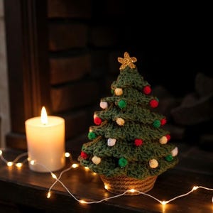 Könnte beinhalten: Ein handgehäkelter grüner Weihnachtsbaum mit roten, weißen und grünen Pompons und einem goldenen Stern. Eine brennende weiße Kerze und Lichterketten neben dem Baum schaffen eine festliche Szene.