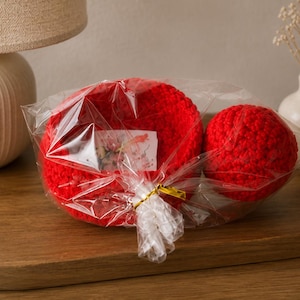 May include: A bright red crocheted bowl and a matching red crocheted sphere are packaged together in clear plastic wrap. A small white rectangular tag with a floral print is tucked inside the plastic, secured with a gold-colored twist tie.
