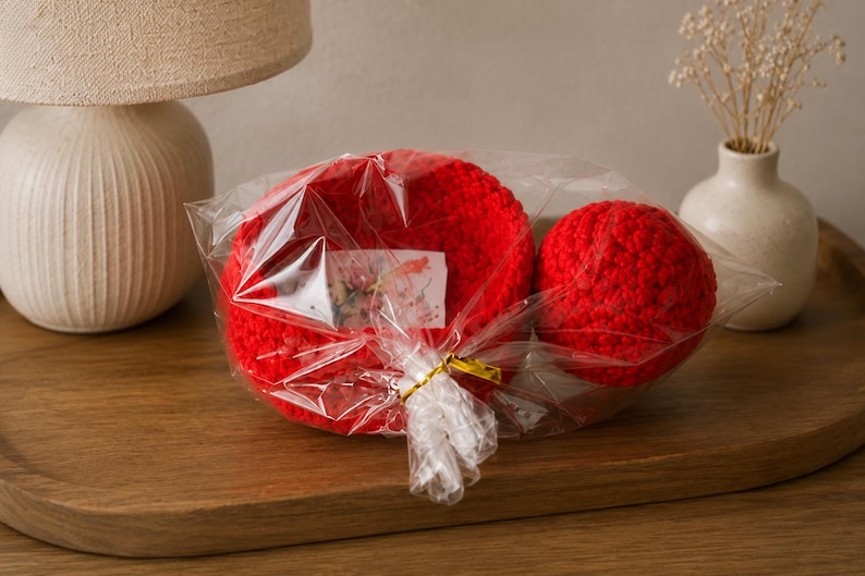 May include: A bright red crocheted bowl and a matching red crocheted sphere are packaged together in clear plastic wrap. A small white rectangular tag with a floral print is tucked inside the plastic, secured with a gold-colored twist tie.