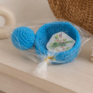 May include: A bright blue crocheted bowl and a matching blue crocheted sphere packaged together in clear plastic wrap. Inside the bowl is a small white card featuring a printed illustration of green asparagus stalks and pink flowers.