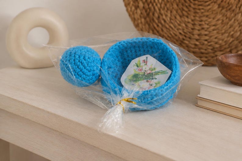 May include: A bright blue crocheted bowl and a matching blue crocheted sphere packaged together in clear plastic wrap. Inside the bowl is a small white card featuring a printed illustration of green asparagus stalks and pink flowers.