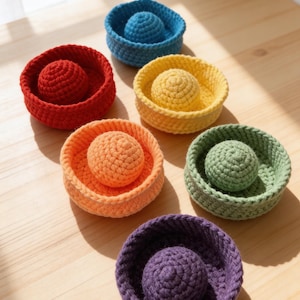 May include: A collection of six circular, crocheted bowls in vibrant colors including red, blue, yellow, orange, green, and purple. Each bowl contains a matching crocheted sphere centered inside, featuring a textured, hand-stitched yarn construction.