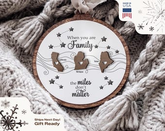 May include: A round wooden ornament featuring a white center with the text When you are Family the miles don't matter. It includes three dark wood cutouts of California, Michigan, and Maine, each with a small heart cutout, connected by lines.