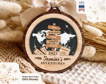 May include: A round wooden ornament featuring a world map graphic, a directional signpost with city names, airplane silhouettes, and travel-themed icons. The text reads 2025, Jasmine's ADVENTURES, Roma, Rio de Janeiro, Dubai, Estambul, and El Cairo.