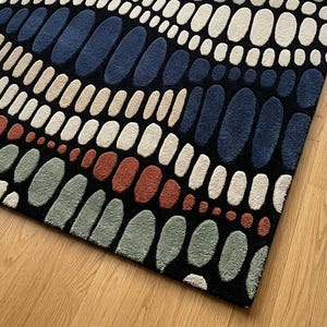 K&ouml;nnte beinhalten: Rechteckiger Teppich mit einem abstrakten Muster aus ovalen Formen in Blau, Beige, Gr&uuml;n und Rostrot auf schwarzem Hintergrund. Der Teppich liegt auf einem Holzboden.
