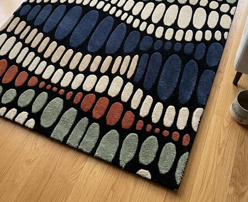K&ouml;nnte beinhalten: Rechteckiger Teppich mit einem abstrakten Muster aus ovalen Formen in Blau, Beige, Gr&uuml;n und Rostrot auf schwarzem Hintergrund. Der Teppich liegt auf einem Holzboden.