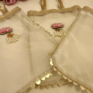 May include: Off-white sheer fabric tote bags with gold trim and handles. Each bag features a pink lotus flower applique with pearl accents. The bags are decorated with gold sequins along the edges. A bouquet of pink roses is in the background.