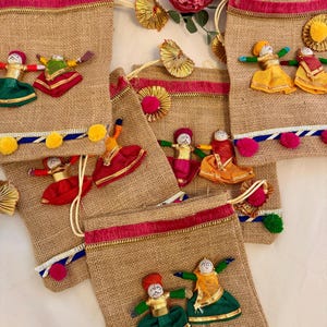 May include: Several burlap drawstring bags decorated with colorful fabric dolls in traditional attire. The bags have a red and pink trim, and are adorned with pom-poms and gold accents. The dolls are dressed in vibrant colors like green, red, and yellow.