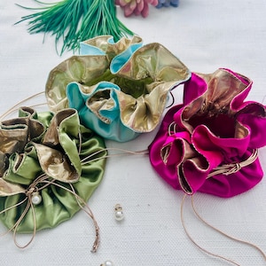 May include: Three drawstring pouches in light blue, olive green, and vibrant magenta. Each pouch features a metallic gold interior lining, a ruffled top edge, and thin gold cord drawstrings finished with small white pearl-like beads.