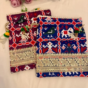 May include: Two decorative pouches with intricate patterns. One is deep purple, the other is blue, both with red and white geometric designs. Gold trim and beaded tassels add detail. Floral arrangement in the background.