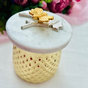 May include: A gold metal container with a textured surface and a white marble lid. The lid features a silver butterfly with gold accents.
