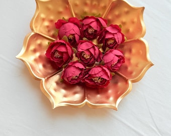 May include: A gold-colored, lotus-shaped decorative tray holds a cluster of deep red artificial flowers. The tray has a metallic finish and is set against a white background, creating a striking contrast. The flowers are arranged in the center of the tray.