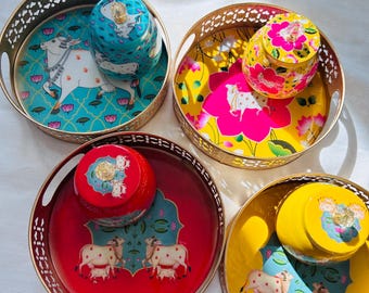 May include: Four round metal trays with gold trim and intricate designs. Each tray features a different color background: turquoise, pink, red, and yellow. Each tray has a small round container with a lid, and each container has a different color: turquoise, yellow, red, and yellow. Each tray features a white cow with a calf, and each tray has a different floral design.