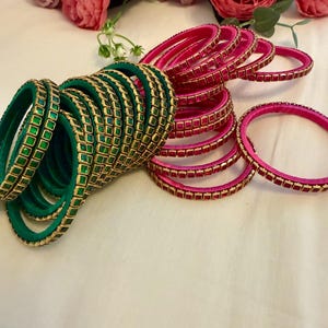 May include: A collection of green and pink bangles with gold-colored accents. The green bangles have a square pattern, while the pink ones have a rectangular pattern. The bangles are arranged on a white surface, with pink flowers in the background.