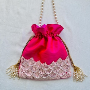 May include: A pink drawstring bag with a pearl and gold chain handle. The bag is decorated with a white pearl trim and has gold tassels.