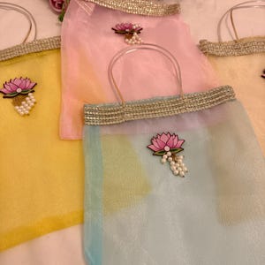 May include: A collection of sheer, colorful tote bags. Each bag features a lotus flower applique with pearl accents and a decorative gold trim. The bags have clear handles and are in shades of yellow, pink, and blue.