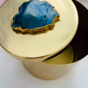May include: A gold-colored metal container with a lid. The lid is adorned with a blue and gold agate stone. The container has a round shape with a slightly textured surface.