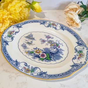 May include: Oval serving platter with a blue and white floral design. The platter features a central scene with a boat and vase of flowers, surrounded by a blue border with floral accents and a yellow trim.