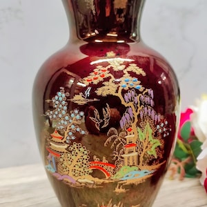 May include: A maroon ceramic vase with a gold rim and a detailed landscape design. The landscape features pagodas, trees, and a bridge, all in gold, red, blue, and green hues. The vase has a glossy finish.