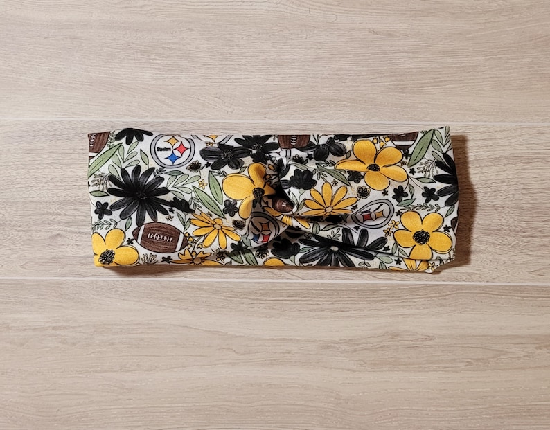 May include: A white headband with a floral pattern featuring yellow flowers, black leaves, and footballs with the Pittsburgh Steelers logo.