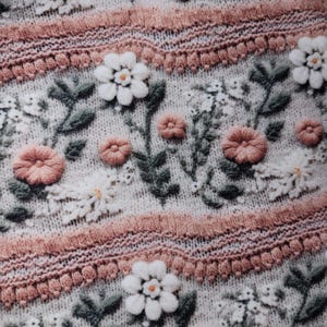 May include: Close-up of a knitted fabric with an embroidered floral design. The pattern features white and peach-colored flowers, green leaves, and decorative pink bands. The fabric has a textured, off-white background.