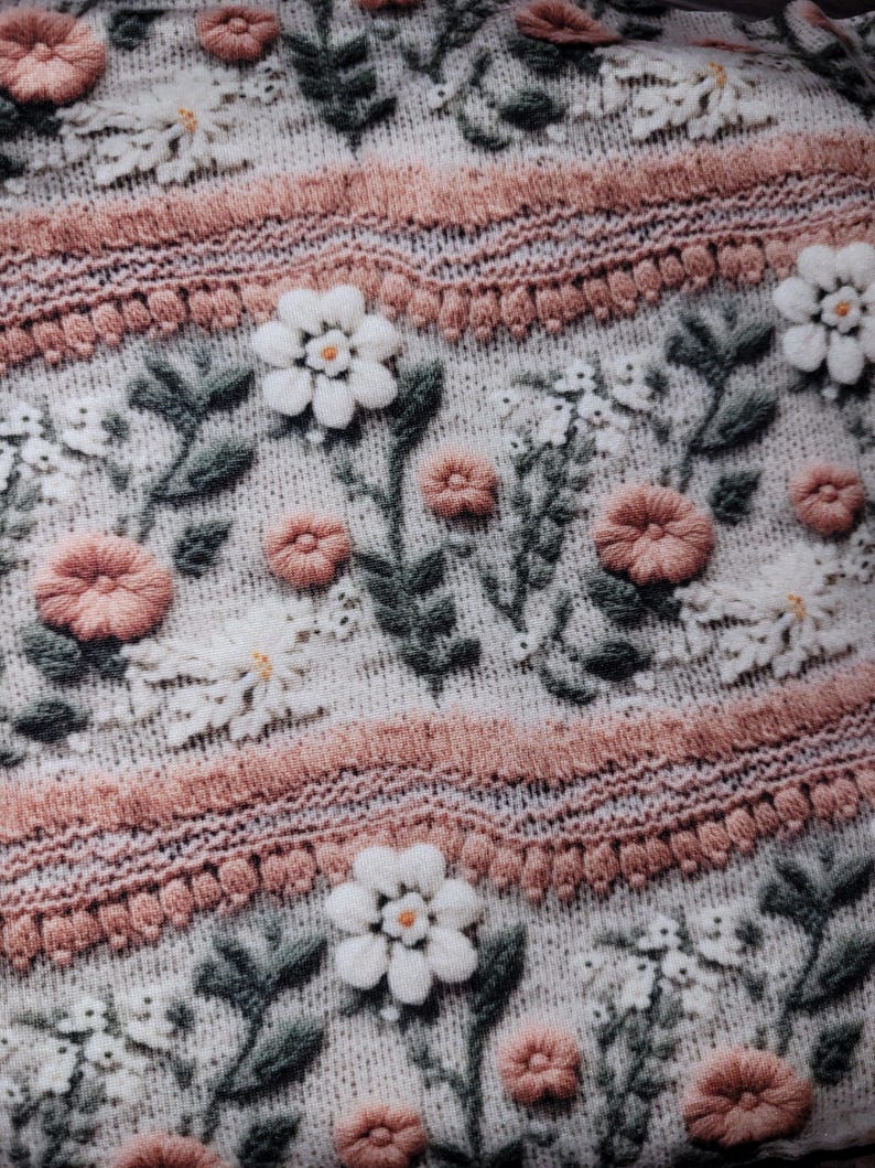 May include: Close-up of a knitted fabric with an embroidered floral design. The pattern features white and peach-colored flowers, green leaves, and decorative pink bands. The fabric has a textured, off-white background.