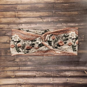 May include: A floral headband with a twisted knot design. The headband features a cream-colored base with a pattern of flowers in shades of pink, white, and green. The edges are trimmed with a textured, light brown knit.