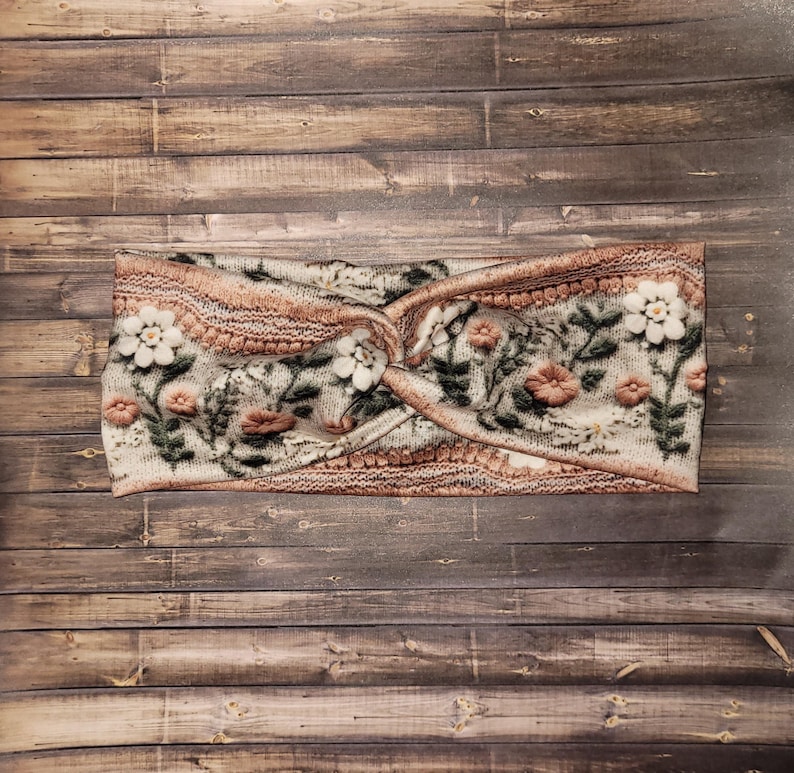 May include: A floral headband with a twisted knot design. The headband features a cream-colored base with a pattern of flowers in shades of pink, white, and green. The edges are trimmed with a textured, light brown knit.