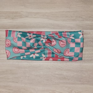 May include: A turquoise and pink headband with a twisted knot design. The headband features a checkerboard pattern, smiley faces, and floral accents. The accessory is designed for comfort and style.