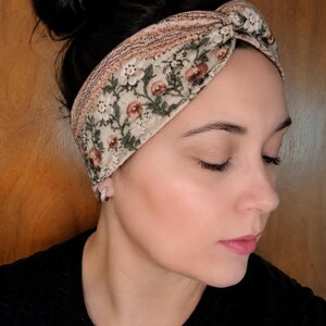 May include: A floral patterned headband in cream, brown, and green. The headband features a knotted design and is worn across the forehead. The floral pattern includes small flowers and leaves. The background is a wooden surface.