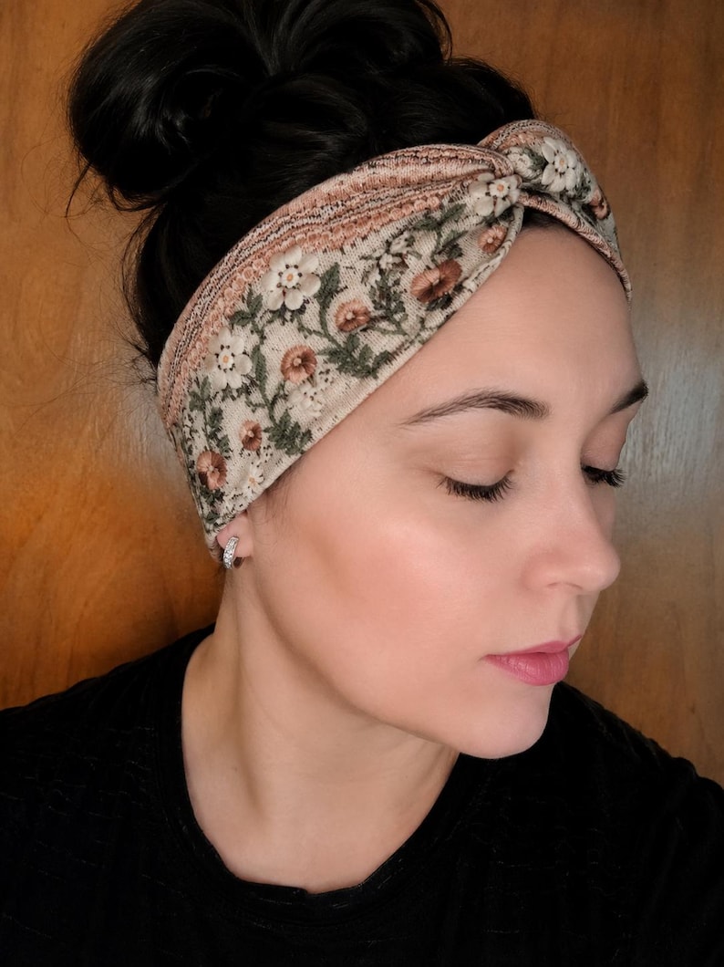 May include: A floral patterned headband in cream, brown, and green. The headband features a knotted design and is worn across the forehead. The floral pattern includes small flowers and leaves. The background is a wooden surface.