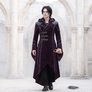 May include: A deep purple velvet coat featuring a hood, structured shoulders, and three horizontal buckle closures on the front. The coat has long, flared sleeves, a high-low hemline, and dark grey trim accents along the seams and shoulders.