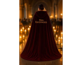 May include: A burgundy velvet hooded cape with the text "Mrs. Ravenwood" embroidered on the hood. The cape is long and flowing, with a deep red color. The background features a dimly lit room with many candles.