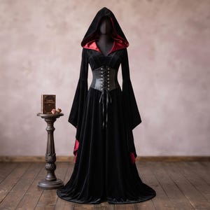 Puede incluir: Un vestido largo de terciopelo negro con capucha, mangas largas acampanadas, forro rojo oscuro y un cinturón tipo corsé negro con ojales plateados y cierre frontal de cordones. La prenda se exhibe sobre un maniquí.