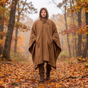 Könnte beinhalten: Eine Person trägt einen braunen Kapuzenponcho mit Knebelverschluss und Taschen. Die Person geht auf einem Pfad durch einen Wald. Der Boden ist mit Herbstlaub bedeckt, und die Bäume haben Herbstlaub.
