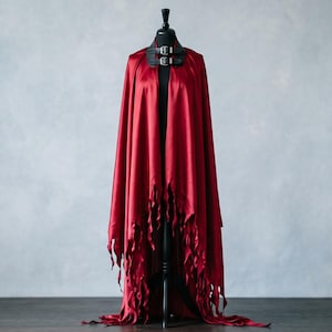 May include: A floor-length, deep red satin cape featuring a high-neck black collar with two silver-toned metal buckles. The hem of the cape is cut into long, thin, jagged strips that hang down to the floor, creating a distressed, layered appearance.