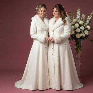 Puede incluir: Dos mujeres con abrigos marfil a juego, largos hasta el suelo, con cuellos y pu&ntilde;os de piel sint&eacute;tica. Los abrigos tienen una botonadura y una falda acampanada. Un gran arreglo floral de rosas blancas est&aacute; en el fondo.