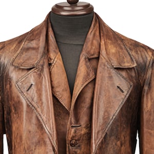 K&ouml;nnte beinhalten: Ein brauner Ledertrenchcoat mit klassischem Design. Der Mantel hat einen Reverskragen, einen Knopfverschluss und eine taillierte Passform. Das Leder hat ein abgenutztes Aussehen, was zu seiner Vintage-&Auml;sthetik beitr&auml;gt. Der Mantel wird auf einer Schaufensterpuppe pr&auml;sentiert.
