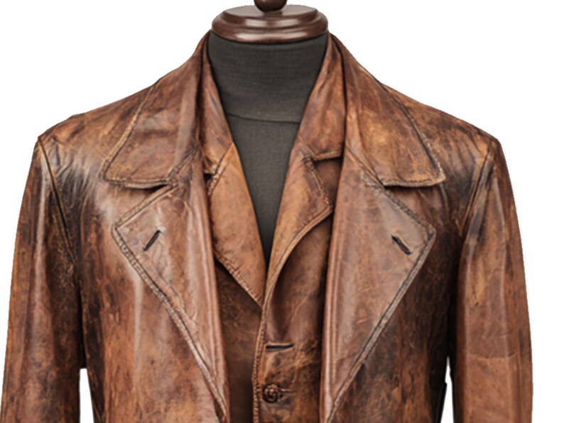 K&ouml;nnte beinhalten: Ein brauner Ledertrenchcoat mit klassischem Design. Der Mantel hat einen Reverskragen, einen Knopfverschluss und eine taillierte Passform. Das Leder hat ein abgenutztes Aussehen, was zu seiner Vintage-&Auml;sthetik beitr&auml;gt. Der Mantel wird auf einer Schaufensterpuppe pr&auml;sentiert.