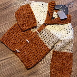May include: A hand-crocheted cardigan in shades of orange, cream, and beige. The cardigan has a button-up front with wooden buttons and a textured stitch pattern. The sleeves and body are colour-blocked, creating a visually appealing design.